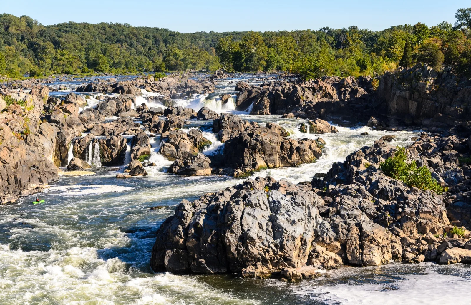 Whitewater rapids pouring over dark rock outcrops between forested banks, a small green kayak in the river